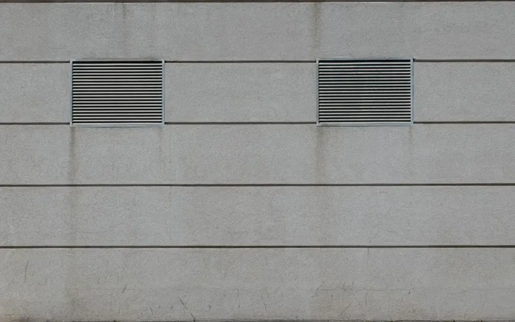 Fasad beton minimalis dengan dua ventilasi persegi sebagai lubang angin roster pada dinding bangunan